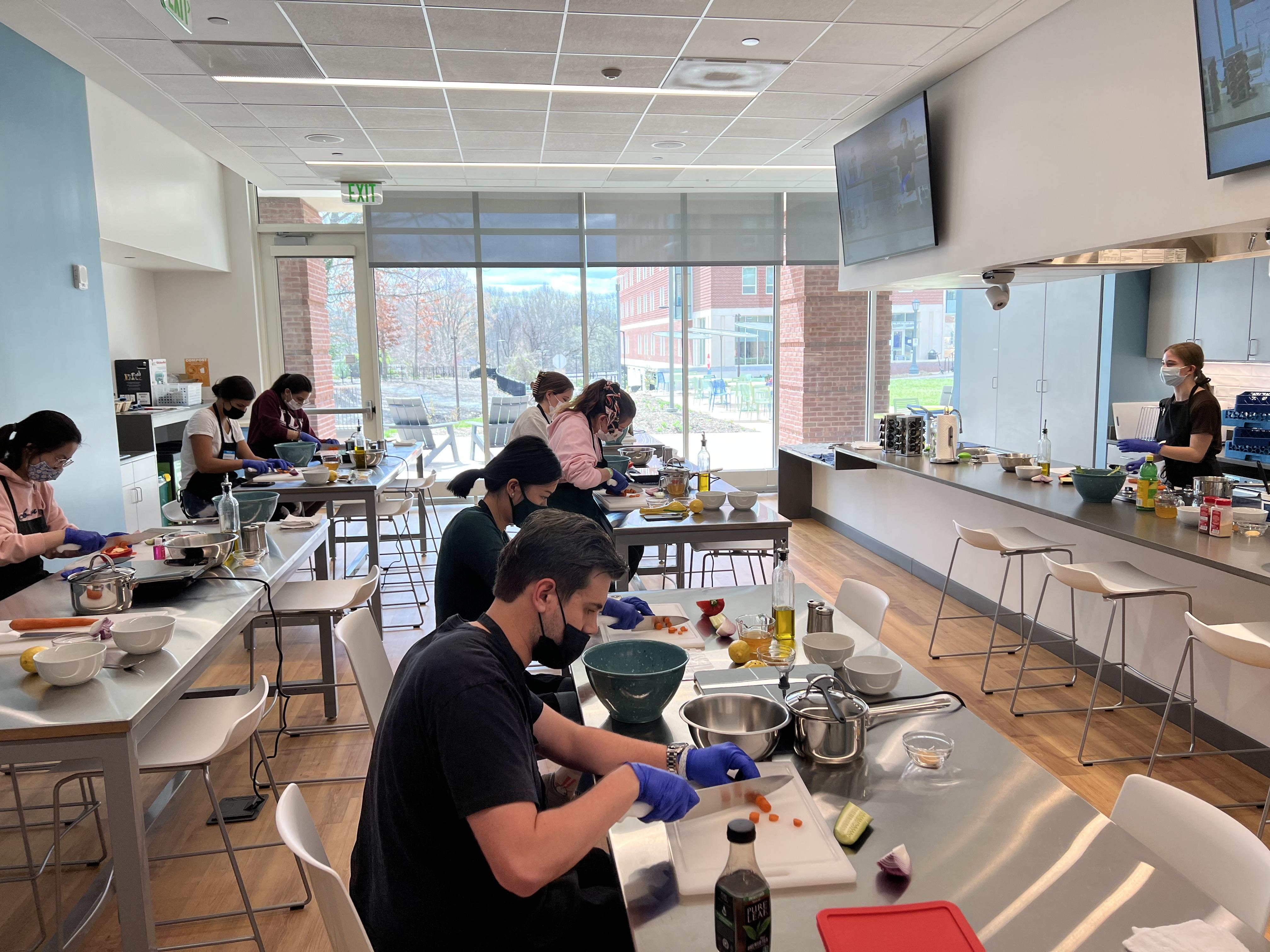 A group of students sits in a teaching kitchen and chops vegetables.