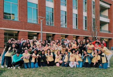 A group of student Peer Health Educators wearing matching yellow t-shirts stand in front of the Student Health and Wellness building and form hearts with their hands.