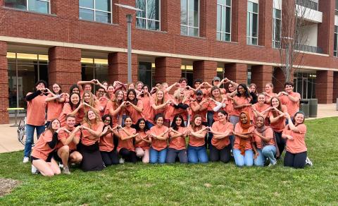 A group of student Peer Health Educators wearing matching yellow t-shirts stand in front of the Student Health and Wellness building and form hearts with their hands.