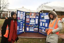 Two women wearing cold weather gear stand in front of a pasteboard outline Peer Health Educator activities and outreach programs.
