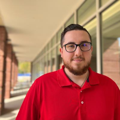Matthew standing outside of the Student Health and Wellness Building wearing glasses and a red polo